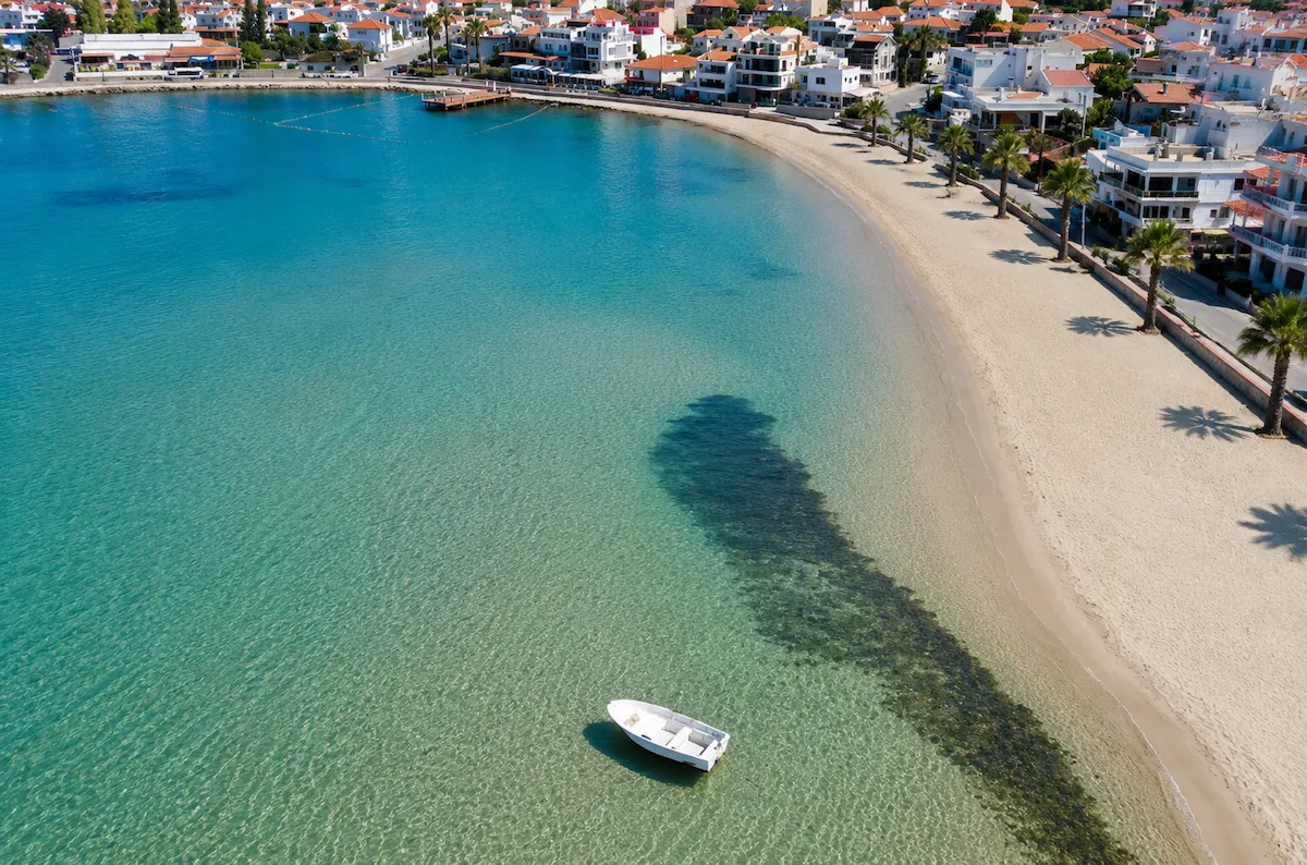 Koca Kum Koyu, İzmir'den çekilmiş bu havadan görünüm, turkuaz renkli berrak denizin, altın kumlu uzun bir plajla birleştiği, kıyı boyunca sıralanmış beyaz evlerin ve palmiye ağaçlarının eşlik ettiği huzurlu bir sahil şeridini gözler önüne seriyor; denizde tek bir beyaz tekne sakin bir şekilde demirlemiş duruyor.