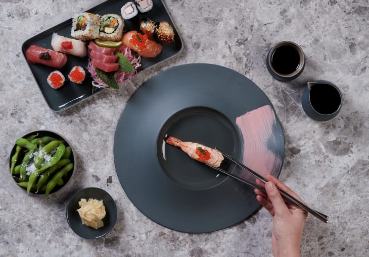 Mermer bir zeminde, bir elin zarifçe chopsticklerle karides nigiriyi aldığı, farklı türde sushi, sashimi, tuzlu edamame ve turşu zencefilin şık koyu gri tabaklar içerisinde sunulduğu bu görsel, İstanbul’un popüler sushi restoranlarında tadabileceğiniz taze ve rafine Uzak Doğu lezzetlerini vurguluyor.