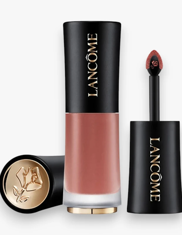 Beyaz bir arka plan üzerinde, şeftali-kahve tonlarındaki Lancôme L'Absolu Rouge Drama Shine likit rujun, altın detaylı, şeffaf alt kısımlı ve üzerinde Lancôme yazısı bulunan dik şişesi, ayrı olarak yatık duran altın gül logolu kapağı ve rujla kaplanmış aplikatörü gösterilmektedir.