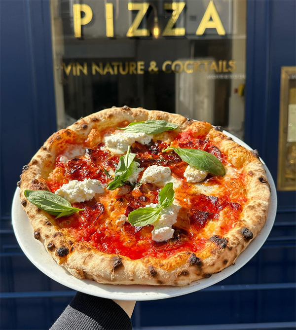 Taze domates sosu, erimiş mozzarella peyniri, beyaz ricotta topları ve taze fesleğen yapraklarıyla dolu, kenarları hafifçe kömürleşmiş, çıtır çıtır bir İtalyan pizzası, arka planda mavi bir vitrin camında En İyi Paris Restoranları arasında yer alan Louie Louie'nin doğal şarap ve kokteyllerini işaret eden altın renkli PIZZA yazısının önünde sunuluyor.
