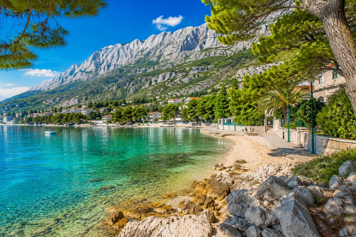 Hırvatistan'daki Makarska Rivierası'nın berrak turkuaz suları, ön planda kayalık kumsalı ve yeşil ağaçlarla süslenmiş kıyı şeridi boyunca uzanan evleriyle, arkasında heybetli Biokovo Dağları'nın yükseldiği güneşli bir günü yansıtan doğal güzelliği.