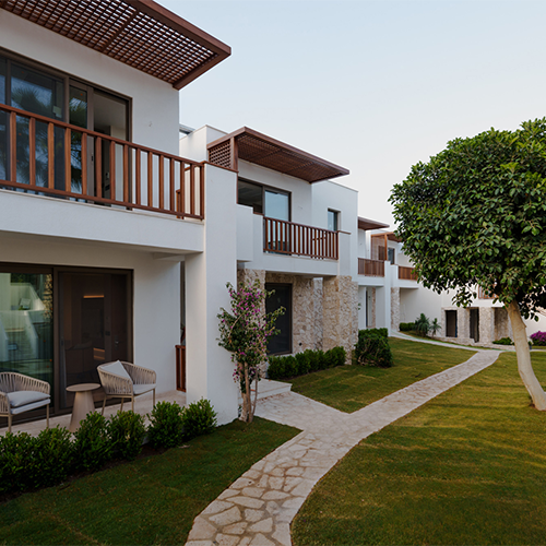 Bodrum'un en iyi otellerinden birini yansıtan bu karede, doğal taş ve ahşap detaylarla bezeli modern beyaz binalar, bakımlı yemyeşil çimler ve kıvrımlı taş yürüyüş yollarıyla çevrili, misafirlere huzurlu bir konaklama deneyimi vadeden şık bir tesis görülüyor.