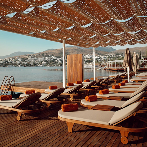 Bodrum'un en iyi otellerinden birinde, deniz kenarında sıralanmış şık bej şezlonglar, üzerinde turuncu havlularla ahşap bir iskelede, tepesinde lazer kesim desenli bronz bir gölgelik ve uzakta Bodrum'a özgü beyaz evlerin bulunduğu tepelerin manzarasıyla lüks ve huzurlu bir atmosfer sunuluyor.