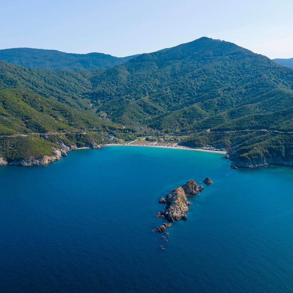 Yemyeşil ormanlarla kaplı heybetli dağların masmavi denize dik indiği, ortasında küçük kayalıkların yükseldiği ve kıyı şeridinde altın kumlu bir plajın uzandığı, kıvrımlı bir yolun dağ yamacını takip ettiği büyüleyici bir koyun kuşbakışı görüntüsü.