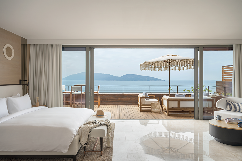 Bodrum'un en iyi otellerinden birinde konumlanan, geniş sürgülü cam kapılarıyla Ege Denizi'nin ve karşıdaki dağların nefes kesen manzarasına açılan, üzerinde şemsiye ve şık oturma grupları bulunan ahşap teraslı, modern ve ferah bir otel odası huzurlu bir tatil vaat ediyor.