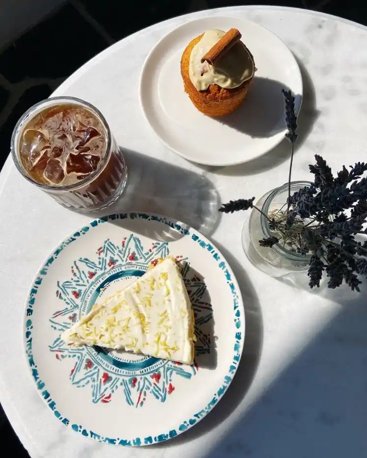 Güneşli bir Ayvalık sabahında, mermer bir masanın üzerinde serinletici bir buzlu kahve, üzerine limon kabukları serpilmiş kremalı bir dilim cheesecake, tarçınlı bir kek ve lavanta dalları, Kuzey Ege'nin keyifli anlarını yansıtıyor.