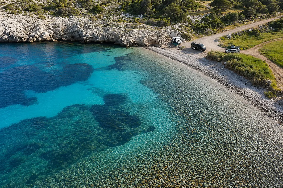Yüksekten çekilmiş bu nefes kesici görüntüde, Karaburun'daki Olcabuk Koyu'nun turkuaz ve lacivert tonlarındaki berrak suları, dibindeki çakıl taşlarını gözler önüne seriyor; kıvrımlı çakıllı sahilde ve toprak yolda park etmiş kampçı ve arazi araçları, bu doğal güzelliğin keşfedilmeyi beklediğini gösteriyor.