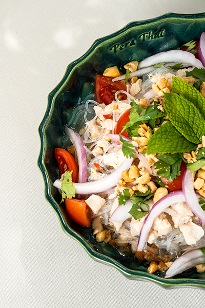 Bodrum'un en iyi restoranları arasında öne çıkan Pera Thai'den gelen bu Tayland usulü noodle salatası, yeşil kenarlı bir kasenin içinde; üzerine taze nane, doğranmış fıstık, kırmızı soğan, domates ve tavuk parçaları eklenmiş şeffaf noodle'lar ile sunularak, otantik lezzetleri göz alıcı bir dengeyle birleştiriyor.