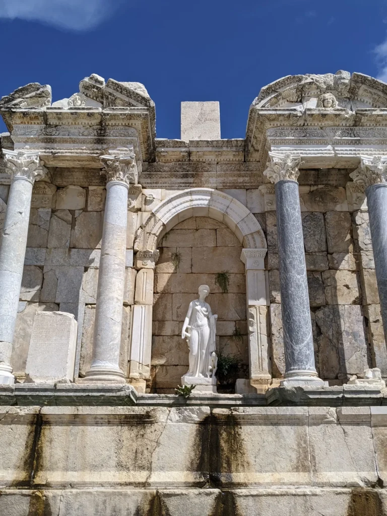 Sagalassos antik kentinden günümüze ulaşan, Roma döneminin ihtişamını yansıtan ve hâlâ su izleri taşıyan Antoninler Çeşmesi'nin merkezindeki zarif beyaz mermer kadın heykeli, çevresindeki gösterişli sütunlar ve detaylı taş işlemelerle süslü yapısıyla, Türkiye'nin antik kentlerinin büyüleyici tarihini gözler önüne seriyor.
