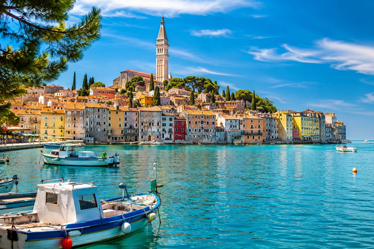 Hırvatistan'ın Rovinj kasabasında, masmavi denizin kıyısında sıralanmış pastel renkli tarihi binalar ve tepesinde yükselen çan kulesi ile pitoresk bir eski şehir sahil manzarası, ön planda demirli teknelerle güneşli bir günde büyüleyici görünüyor.
