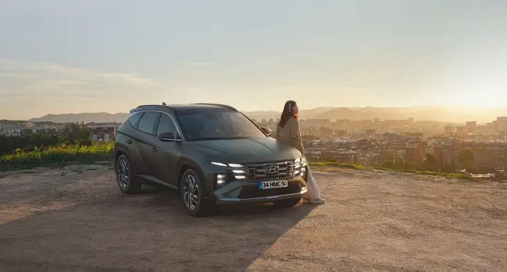 Hyundai Tucson SUV, gün batımında şehir manzarasına karşı park halinde; yanında manzarayı izleyen kadın silüeti.