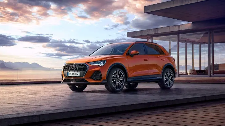 Turuncu Audi Q3 Sportback, modern mimari ve gün batımı manzarası eşliğinde şık ve sportif tasarımıyla sergileniyor.