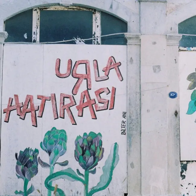 Urla Sanat Sokağı'nda yer alan beyaz bir duvarda, Urla'nın sembolü haline gelmiş enginar çizimleri eşliğinde kırmızı harflerle URLA HATIRASI yazısı ve üstte eski bir pencere detayıyla, bölgenin doğal ve kültürel atmosferi yansıtılıyor.