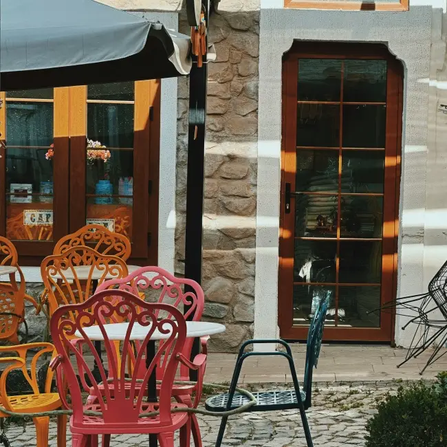 Urla'nın tarihi Sanat Sokağı'nda, rengarenk ferforje sandalyelerin ve yuvarlak masaların kaldırımda sıralandığı, taş dokulu bir binanın ahşap pencerelerinden güneş ışıklarının süzüldüğü davetkar bir kafe köşesi, bu seyahat rehberi için Urla'nın otantik atmosferini yansıtıyor.