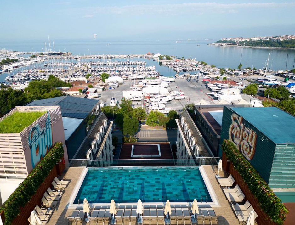 İstanbul'daki Wyndham Grand Kalamış Marina Hotel'in terasından çekilen bu görselde, Kalamış Marina'nın yatlarla dolu hareketli panoramasını kucaklayan cam kenarlı turkuaz bir sonsuzluk havuzu, etrafındaki şezlonglar ve şemsiyelerle şehirde unutulmaz bir yaz kaçamağı vaat ediyor.