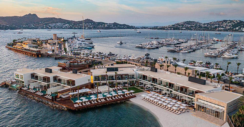 Akşamüstü çekilmiş bu geniş açılı görselde, Yalıkavak Marina'nın berrak sularına nazır konumlanmış lüks bir otel, özel beyaz kumsalı, havuzları ve şezlonglarıyla dikkat çekerken, marina yatlarla dolu ve ardındaki tepelerde de Bodrum'un otelleri ve evleri yer alıyor.