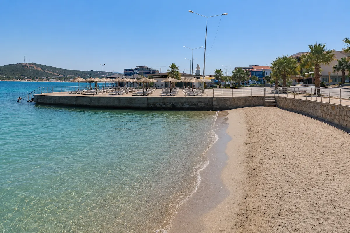 Güneşli bir günde, Çeşme İzmir'deki Yıldızburnu Plajı'nda, berrak turkuaz denizin kıyıya vurduğu kumlu bir plaj ve şemsiyelerle donatılmış iskelenin ardında palmiye ağaçları ile binalar görülüyor, bu bölge aynı zamanda termal plaj özelliğiyle biliniyor.