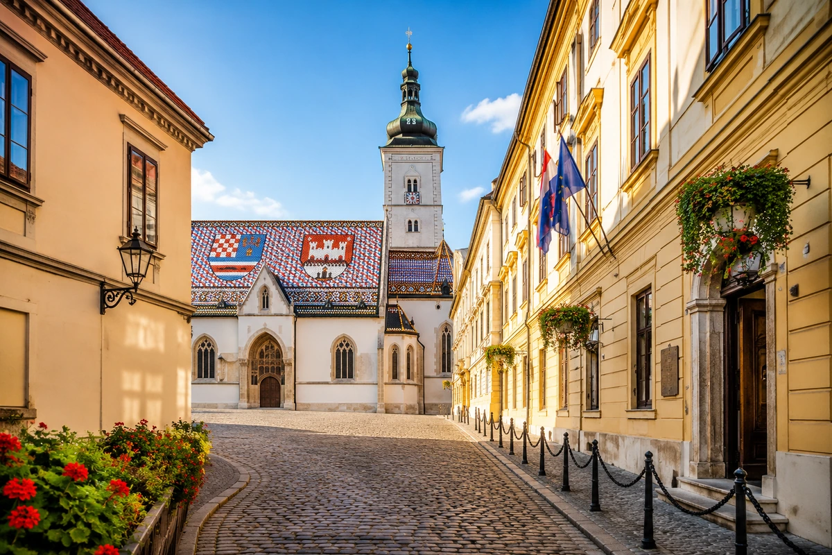 Hırvatistan'ın başkenti Zagreb'deki tarihi St. Mark Kilisesi Sokağı, armalı mozaik çatısıyla öne çıkan St. Mark Kilisesi'nin görkemli kulesi, kırmızı çiçeklerle bezenmiş kaldırım kenarı ve asılı çiçek sepetleriyle süslü sarı binalarla, güneşli bir günde taş döşeli bir manzarayı gözler önüne seriyor.