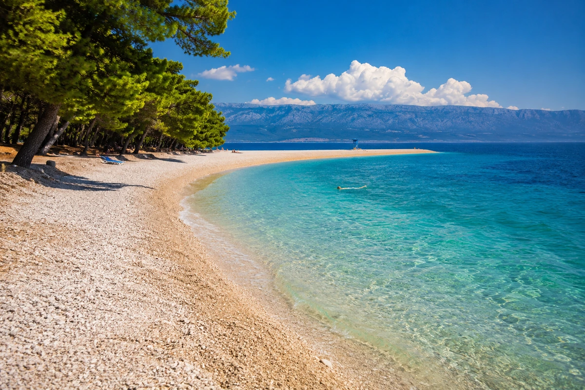 Zlatni Rat Plajı'nda, yemyeşil çam ağaçlarının gölgelediği çakıllı kıyının berrak turkuaz sularla buluştuğu bu eşsiz sahil manzarası, uzakta yüzen bir kişi ve uç kısmında cankurtaran kulesi olan meşhur burun ile canlanıyor.