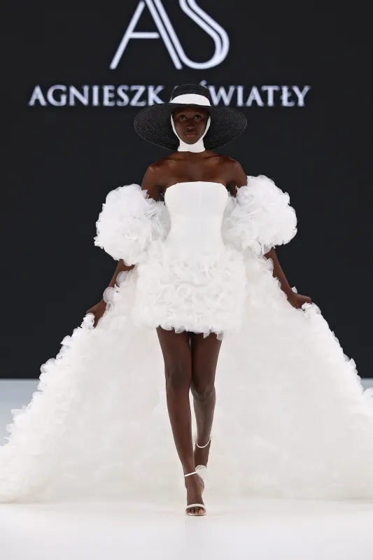 Model, kabarık kolları ve dramatik kuyruğuyla dikkat çeken, mini etek boyunda beyaz bir gelinlik giymekte; geniş siyah-beyaz şapka ve beyaz bant aksesuarla podyumda çarpıcı bir görünüm sergiliyor.