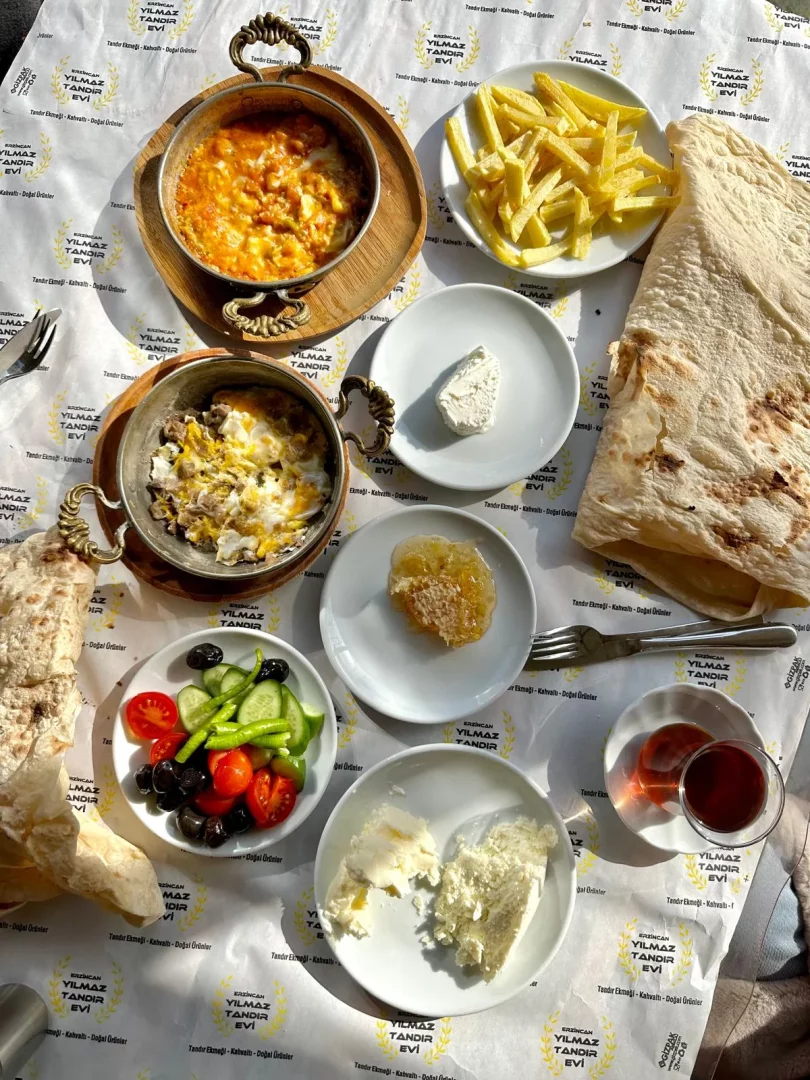 Erzincan Yılmaz Tandır Evi'nin geniş bir kahvaltı menüsü sunan bu görselinde, bakır sahanlarda menemen ve etli yumurta, patates kızartması, bal peteği, zeytin, taze salata, farklı peynir çeşitleri ve tandır ekmeği ile hazırlanan doyurucu bir sofra, güneşli bir ortamda havadan görüntüleniyor.