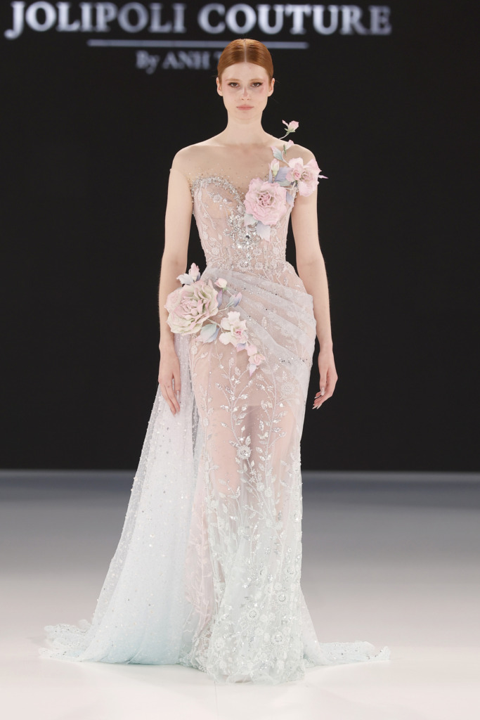 Model, pastel mavi ve pembe tonlarında işlenmiş, transparan ve ışıltılı dokuya sahip tek omuzlu bir gelinlik giyiyor; üzerindeki 3D çiçek aplikeleri romantik bir görünüm yaratıyor.