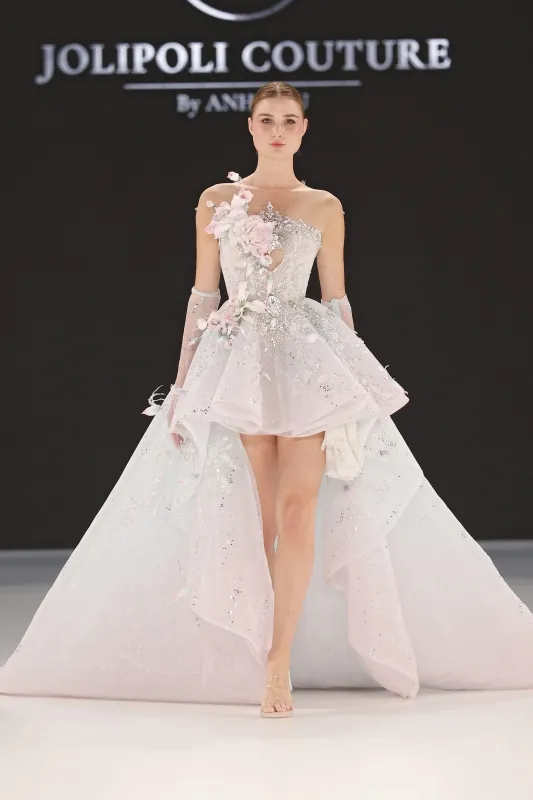 Model, tek omuzlu, ön kısmı mini formda tasarlanmış ve arkası uzun kuyrukla tamamlanan, çiçek aplikeleri ve taş işlemelerle süslenmiş pastel tonlu bir gelinlik giyiyor.
