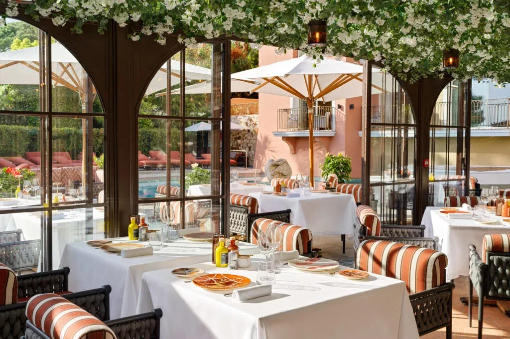 Saint-Tropez'deki Hotel Byblos'un Il Giardino restoranının aydınlık ve lüks yemek alanı, beyaz örtülü masaları, çizgili minderli sandalyeleri, tavandan sarkan yemyeşil çiçekli bitkileri ve camların ardında görünen havuz kenarındaki şezlonglar ile büyük beyaz şemsiyelerle göz alıcı bir atmosfer sunuyor.