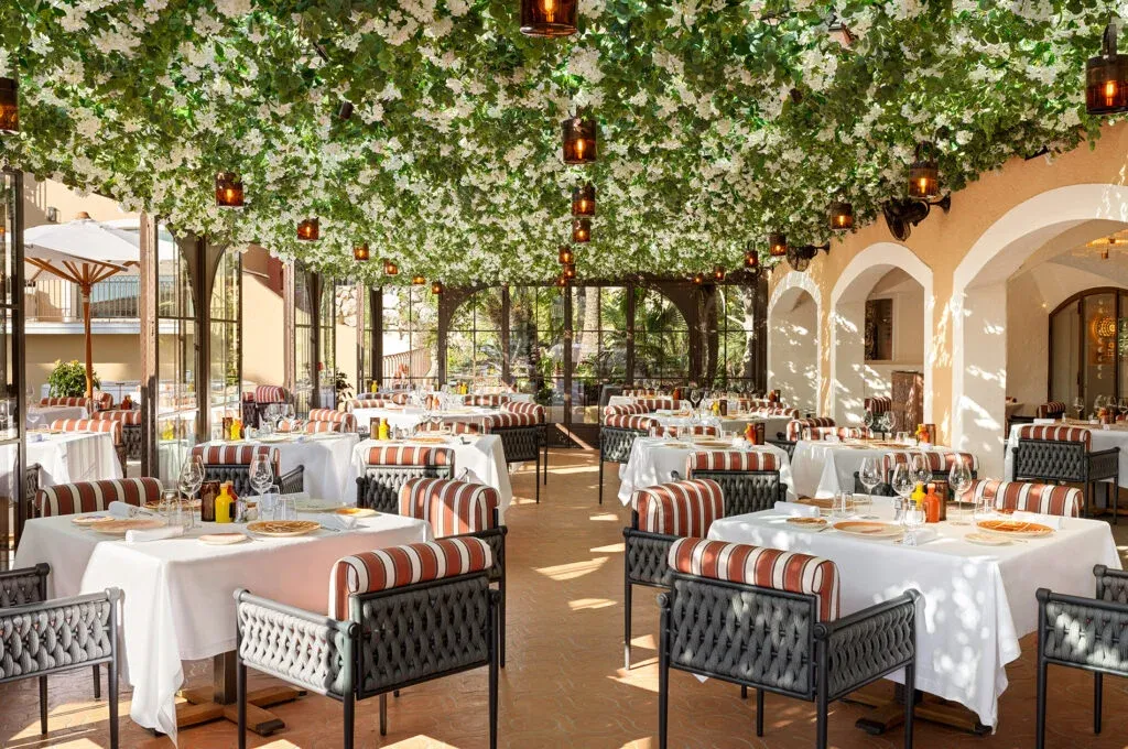 Saint-Tropez'deki Hotel Byblos bünyesinde yer alan Il Giardino restoranında, tavandan sarkan bol beyaz çiçeklerle bezeli yemyeşil yaprakların oluşturduğu doğal bir gölgelik altında, zarif beyaz örtülü masalar ve çizgili minderli sandalyelerle dolu, aydınlık ve davetkar bir yemek alanı görülüyor.