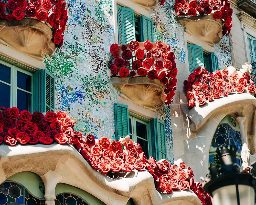 Barselona’daki Casa Batlló’nun renkli mozaik cephe detayı; balkonları çevreleyen kırmızı gül süslemeleri ve yeşil panjurlu pencereler.
