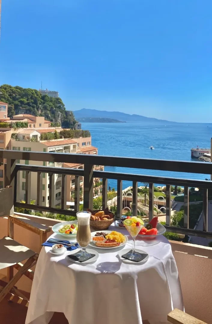 Columbus Otel Monte Carlo'nun Akdeniz manzaralı balkonunda, üzerinde beyaz örtü serili bir masada sunulan zengin kahvaltıda çırpılmış yumurta, sosis, kruvasan, meyve salatası ve köpüklü latte gibi birçok lezzet, masmavi deniz ve yeşil tepelerin eşsiz güzelliğiyle birleşiyor.
