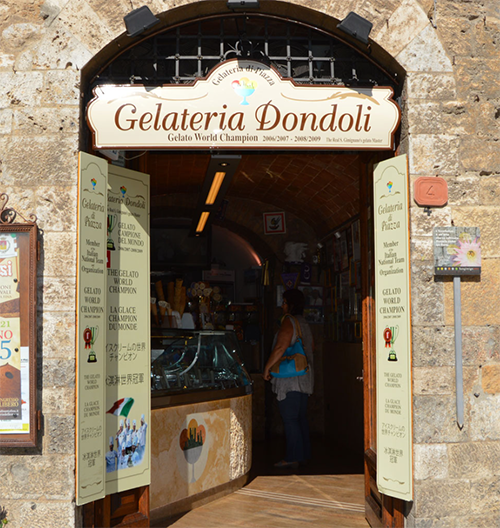 San Gimignano kasabasında, geleneksel kemerli taş girişli binasında Gelateria Dondoli tabelasıyla dünyanın en iyi dondurmacısı unvanını sergileyen dükkanın içinde tezgahın önünde duran bir ziyaretçinin göründüğü bu görüntü, Toskana'nın lezzet duraklarından birine davet ediyor.