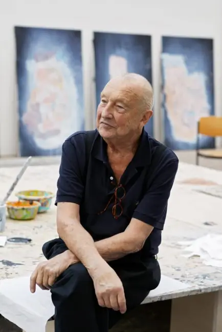 Sanatçı Georg Baselitz, sanat yaşamının önemli bir figürü olarak tanınan, kel, koyu renk polo tişörtlü ve boynunda asılı gözlüklü bir şekilde, arkasında soyut tablolar ve önünde boya kaseleri bulunan atölyesinde oturmuş, yan tarafa doğru düşünceli bir ifadeyle bakıyor.