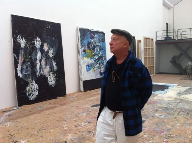 Sanatçı Georg Baselitz, geniş ve boya sıçramış zeminli atölyesinde, karakteristik baş aşağı figürler içeren koyu renkli ve belirgin fırça darbelerine sahip soyut eserlerinden birine dikkatle bakarken gri şapkalı, ekose gömlekli bir şekilde duruyor.