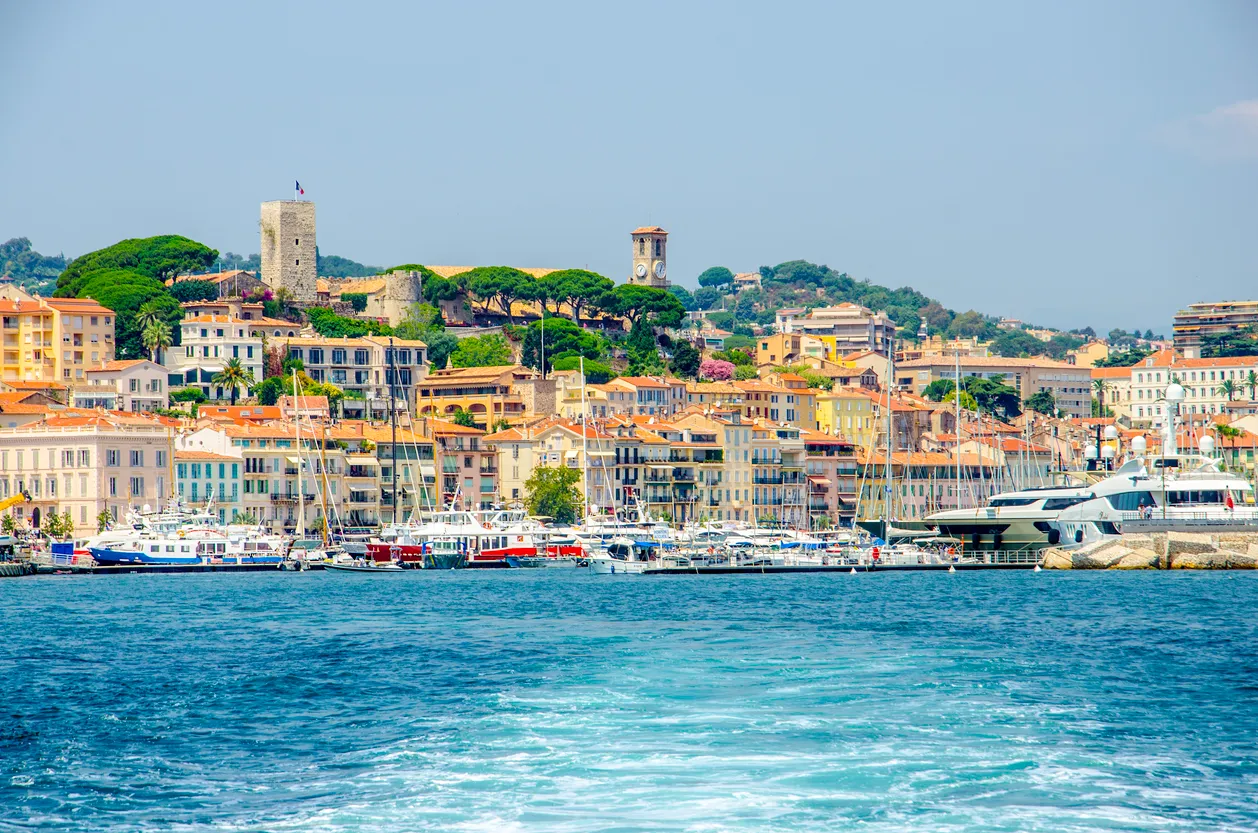 Denizden çekilmiş bu panoramik Güney Fransa görüntüsünde, masmavi sularda ilerlerken sağda ve solda sıralanmış teknelerle dolu bir liman ve tepelere yayılan, farklı renklerdeki binaların ve yeşil ağaçların süslediği canlı bir sahil kasabası görünüyor, kasabanın ortasında bir saat kulesi ve sol tarafta ise tarihi bir taş kule dikkat çekiyor.
