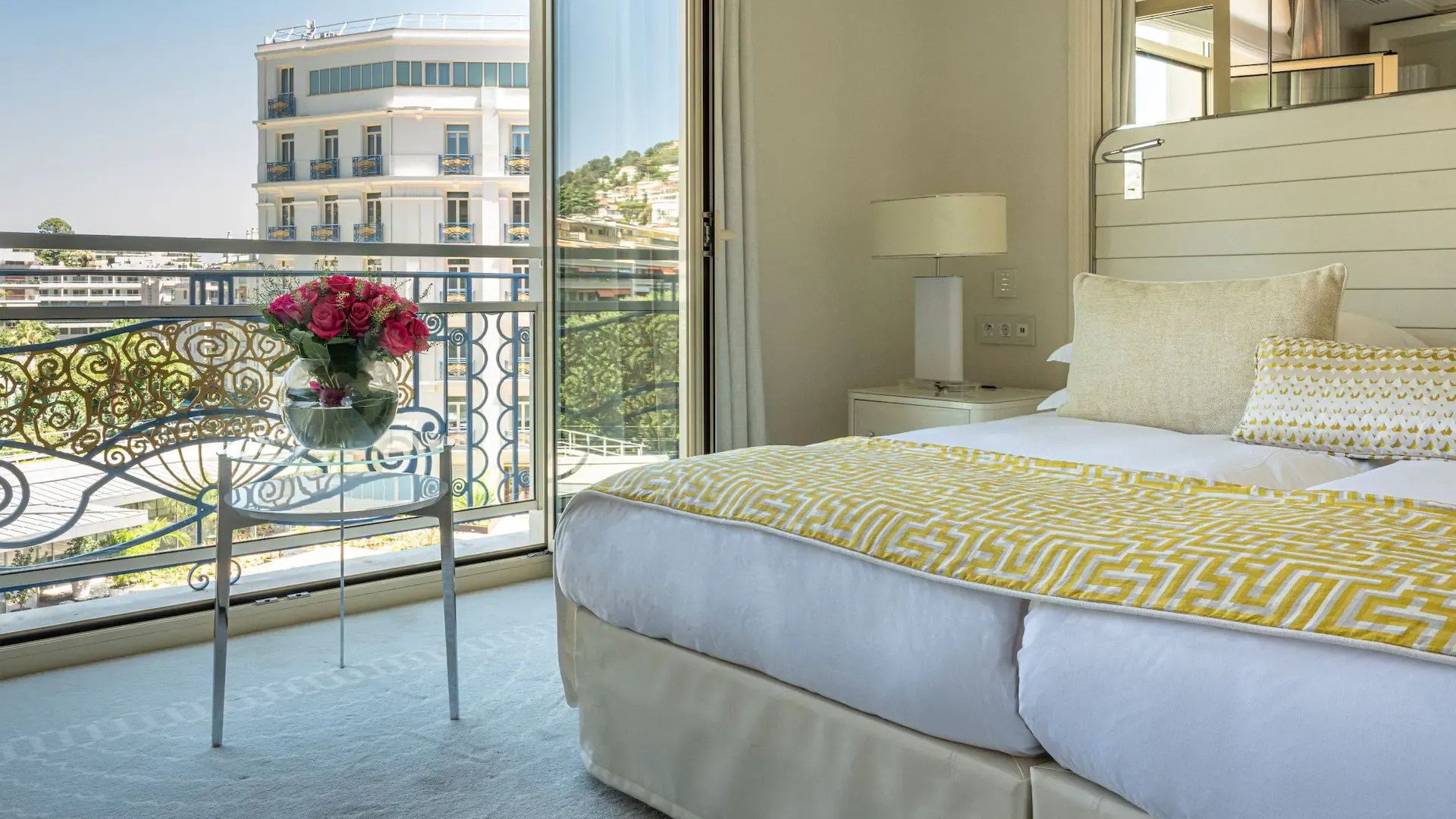 Aydınlık ve lüks bir otel odasının balkonundan, ön planda pembe güllerle dolu şeffaf bir vazonun durduğu cam masa ve oymalı korkuluklar ile çevrili şehir manzarası görülüyor; sarı geometrik desenli pike örtülü yatak ve beyaz modern komodin odanın zarif konforunu tamamlıyor.
