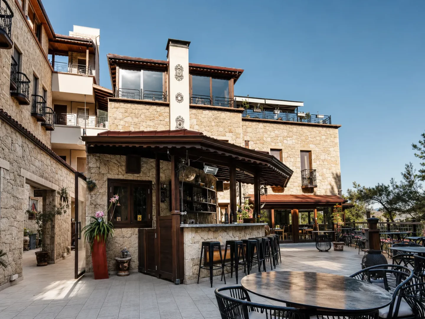 Kaz Dağları'nın doğal güzellikleri arasında yer alan Karye Müze Otel'in taş cepheli binası önündeki geniş terasta, ahşap çatılı şık bir açık hava barı ve misafirler için düzenlenmiş modern oturma grupları güneşli bir havada görülüyor.