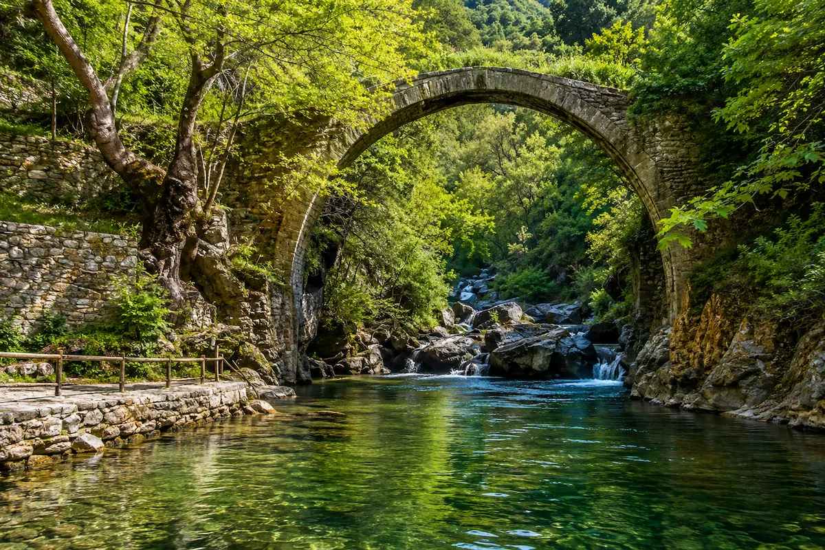 Yeşillikler içinde, taş kemerli tarihi bir köprünün Mihlı Çayı üzerinde yükseldiği, berrak suların aktığı, kenarında ahşap korkuluklu taş bir yolun uzandığı ve ileride küçük şelalelerin görüldüğü görkemli bir doğa manzarası.