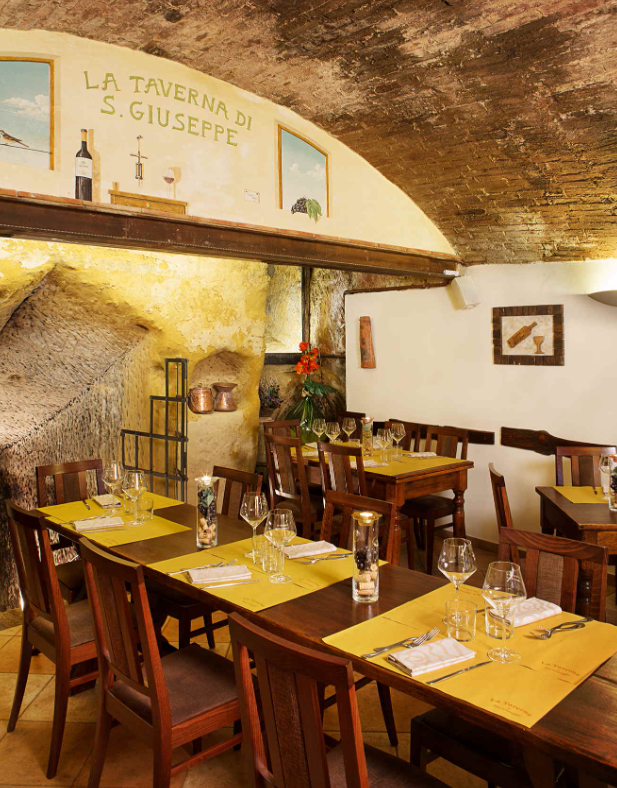 Siena'daki La Taverna di San Giuseppe restoranının kemerli tuğla tavanlı ve taş duvarlı iç mekanında, sarı Amerikan servisli masalar özenle hazırlanmış olup, Toskana'nın kültürel mirasını ve zengin gastronomi deneyimini yansıtan sıcak bir atmosfer sunuyor.