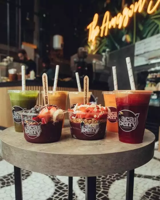 Acai berry markalı şeffaf kaplarda sunulan çeşitli içecekler ve üstü meyveli, granolalı iki büyük açai kasesinin, arka planda Amazon temalı neon bir yazı ve tropikal bitkilerle modern bir kafenin masasında iştah açıcı bir şekilde durduğu, sağlıklı beslenmeyi çağrıştıran çekici bir görüntü.