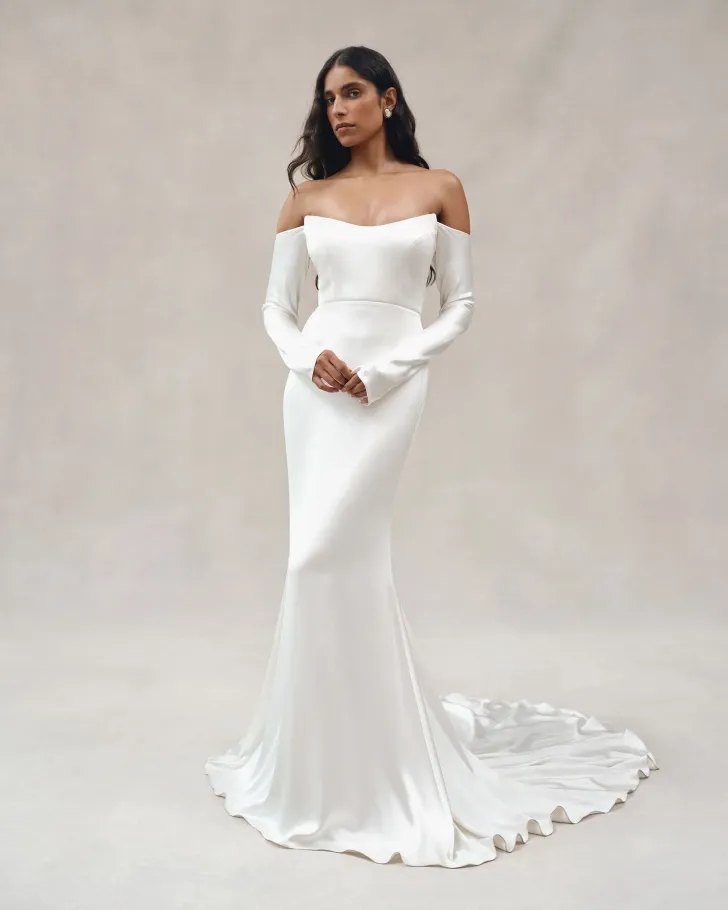Alexandra Grecco tasarımı, düşük omuzlu ve uzun kollu saten gelinlik modeli. Vücuda oturan silueti ve zarif kuyruk detayıyla minimal ama etkileyici bir bridal görünüm sunuyor.