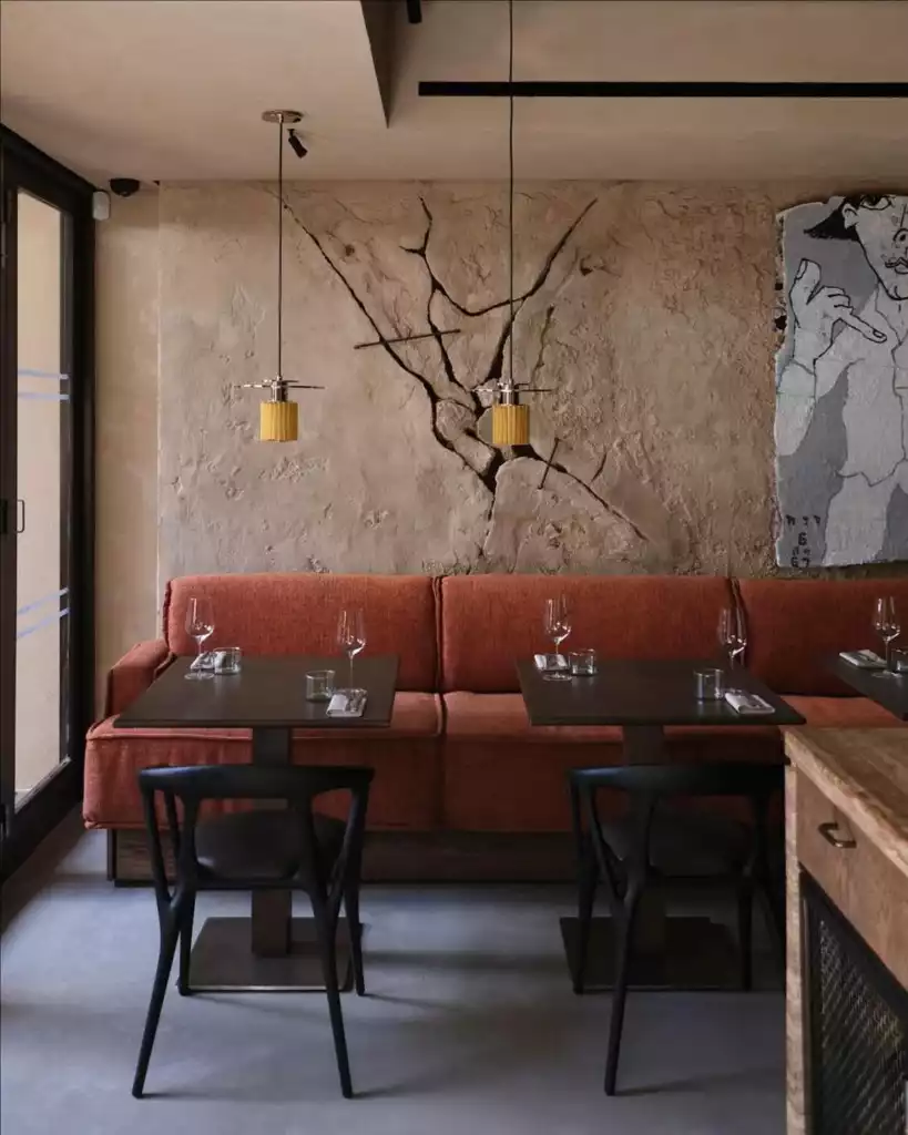 Modern bir restoran iç mekanı, çatlak deseniyle dikkat çeken dokulu açık renkli duvar önünde uzanan turuncu döşemeli bankı, üzerinde kadeh ve peçetelerin bulunduğu iki koyu renkli masayı, siyah sandalyeleri ve tavandan sarkan sarı metalik iki sarkıt lambayı gösteriyor, sağ tarafta ise soyut bir duvar sanatı yer alıyor.