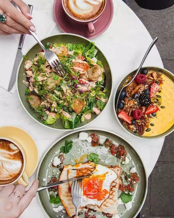 Apple Butter Cafe'deki bir masada, çatalıyla yeşillik, tavuk parçaları ve soslu büyük bir salata yiyen bir el ile akışkan sarılı yumurtalı gözlemeyi kesen başka bir elin görüldüğü, yanında granola ve taze meyvelerle süslenmiş parlak sarı smoothie kasesi ve latte artlı kahvelerle dolu, zengin bir brunch sofrası sergileniyor.