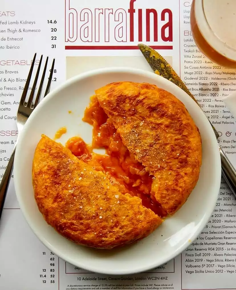 Barrafina restoranının menüsü ve sofradaki çatal bıçak eşliğinde, beyaz bir tabakta duran, ortadan ikiye ayrılmış ve sıcak, akışkan patatesli iç harcı görünen, üzerinde iri tuz taneleri parlayan iştah açıcı bir İspanyol tortilla.
