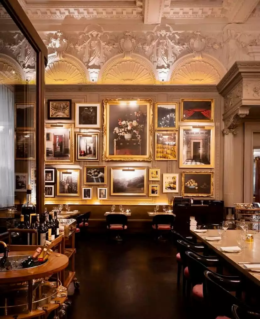 Berners Tavern'ın lüks ve sanatsal atmosferini yansıtan bu iç mekanda, duvarlar tavanlara kadar uzanan heykelsi kabartmaların altında, çiçek resimleri ve manzaralar dahil olmak üzere farklı boyutlarda onlarca altın varaklı çerçeveli tabloyla dolu, loş ışıklandırmalı bir ortamda şık bar ve yemek masaları dikkat çekiyor.