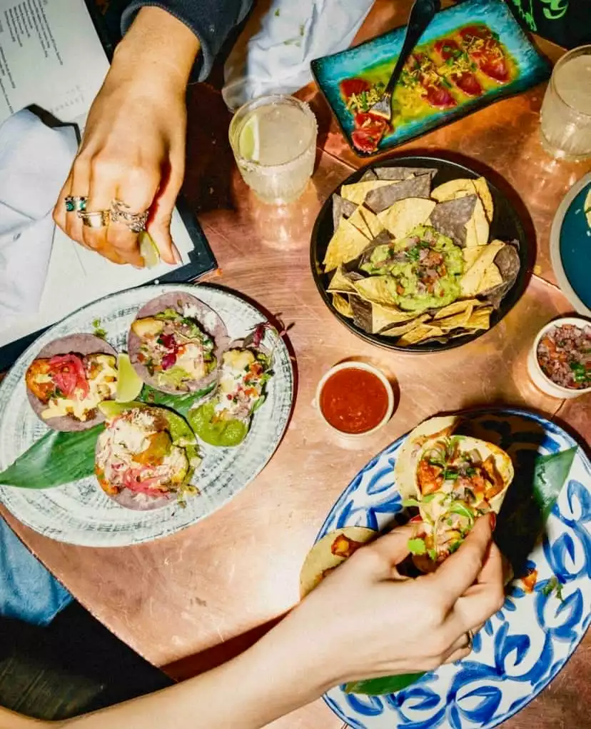 Bodega Negra'da bakır rengi bir masada, iki elin tacoları hazırladığı ve yediği, mor mısır tortillalı tacolar, bol guacamole'li cipsler, taze ceviche ve margarita gibi zengin Meksika mutfağı lezzetlerinin üstten çekim sunumu.
