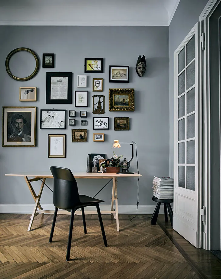 Koyu gri duvarda antika ve modern çerçevelerle oluşturulmuş asimetrik galeri düzeni; retro bir çalışma masasıyla tamamlanan eklektik alan.