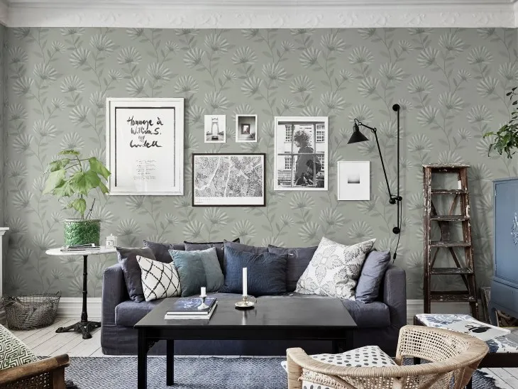 Yaprak desenli duvar kağıdı üzerinde siyah-beyaz çerçeveli fotoğrafların yer aldığı modern salon; mavi kanepe ve karışık dokulu yastıklarla tamamlanmış.