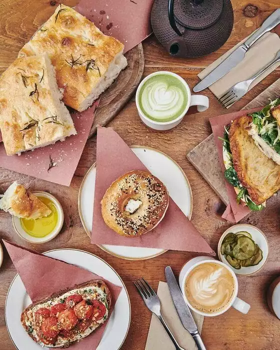 Ahşap bir masada üstten çekilmiş bu fotoğrafta, biberiyeli focaccia ekmekleri, susamlı ve haşhaşlı bagel, domatesli tost, sandviç, zeytinyağı, turşu, kalpli matcha latte ve yapraklı latte gibi çeşitli fırın ürünleri ve kahvaltılıklar özenle sergileniyor.