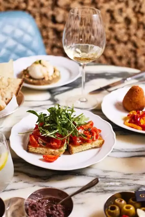 Mermer bir masada, domates ve roka yapraklarıyla zenginleştirilmiş iki dilim bruschetta, bir kadeh beyaz şarap, zeytin, zeytin ezmesi, altın rengi kızarmış arancini ve üzerinde peynir topu ile fıstıklar bulunan bir başka meze gibi çeşitli iştah açıcı İtalyan Cicchetti ve aperatifleri sergileniyor.